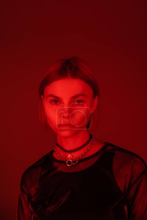 portrait of young and pretty woman in necklaces looking at camera in red light on burgundy background