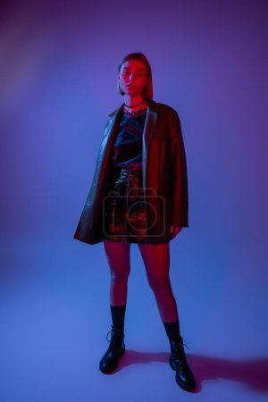 full length of young woman in leather jacket with mini skirt and black boots standing on blue background