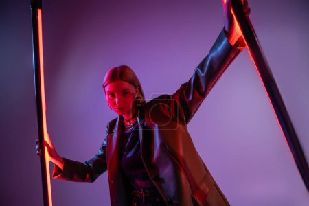 Photo for Fashionable woman in leather jacket looking at camera near bright neon lamps on purple background - Royalty Free Image