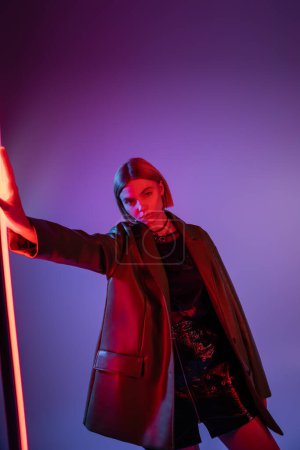 young woman in mini skirt and leather jacket standing near vibrant neon lamp on purple background