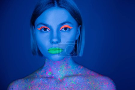 retrato de mujer en maquillaje de neón vibrante y pintura corporal fluorescente mirando a la cámara aislada en azul oscuro