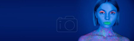 Foto de Retrato de mujer con labios de neón verde y sombra de ojos naranja mirando a la cámara aislada en azul oscuro, pancarta - Imagen libre de derechos