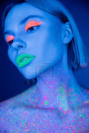 portrait of young woman with fluorescent makeup and neon paint on body isolated on dark blue