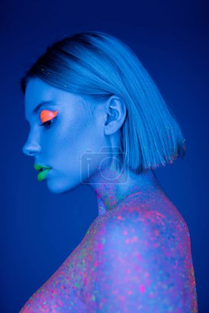 Foto de Perfil de mujer joven con maquillaje de neón y salpicaduras de pintura brillante sobre fondo azul - Imagen libre de derechos