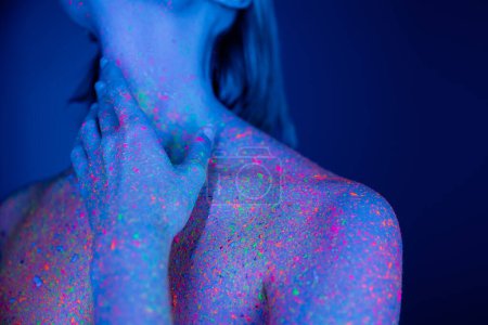 vue recadrée de femme nue colorée en peinture au néon brillant touchant le cou sur fond bleu 
