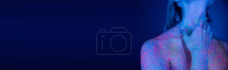 Téléchargez les photos : Vue recadrée de femme nue avec éclaboussures de néon lumineux sur le corps touchant le cou sur fond bleu foncé, bannière - en image libre de droit