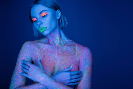 naked woman in neon makeup and bright paint splashes covering bust with hands isolated on dark blue