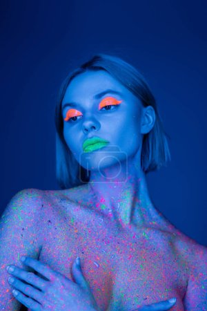 Foto de Mujer desnuda en maquillaje fluorescente y pintura corporal de neón que cubre el pecho con las manos aisladas en azul oscuro - Imagen libre de derechos