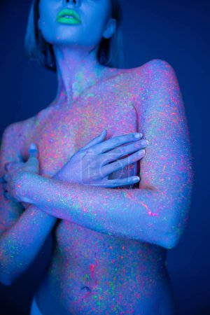 Teilansicht einer nackten Frau mit grünen Neon-Lippen und farbenfroher Körperfarbe, die die Brust bedeckt, mit isolierten Händen auf dunkelblau