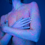 partial view of nude woman with green neon lips and colorful body paint covering breast with hands isolated on dark blue