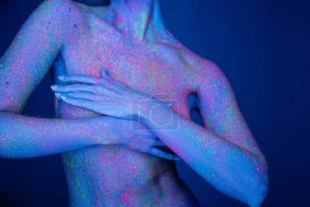 Ausgeschnittene Ansicht einer nackten Frau mit hellen Neon-Spritzern auf der Brust, die den Körper bedeckt, mit isolierten Händen auf dunkelblau