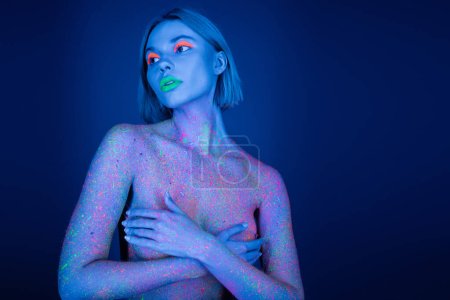 naked woman in neon makeup and bright paint splashes covering bust with hands and looking away isolated on dark blue