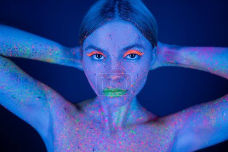 retrato de mujer con maquillaje de neón vibrante y salpicaduras de pintura brillante en el cuerpo sosteniendo las manos detrás de la cabeza aislado en azul oscuro