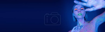 Téléchargez les photos : Femme au néon vibrant peinture corporelle et maquillage posant avec la main tendue floue sur fond bleu foncé, bannière - en image libre de droit