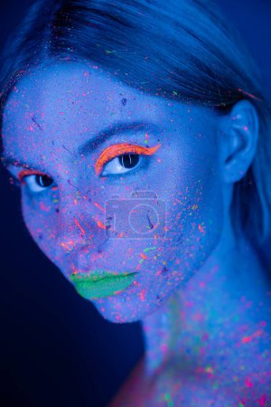 Téléchargez les photos : Portrait de femme avec maquillage fluo et peinture fluorescente sur le visage isolé sur bleu foncé - en image libre de droit