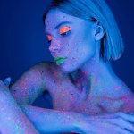 nude woman with neon makeup and body in fluorescent paint isolated on dark blue