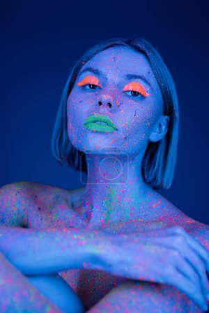 Porträt einer jungen Frau in grellem Neon-Make-up und farbenfroher Körperfarbe, die isoliert auf dunkelblau in die Kamera blickt