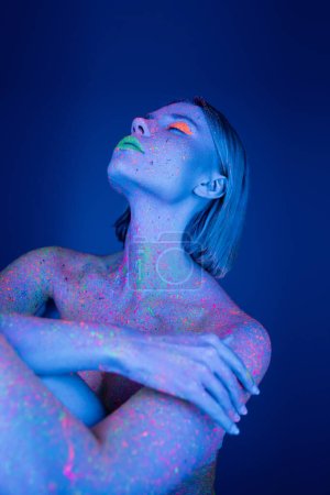 nackte Frau in hellem Make-up und Neon-Körperfarbe posiert auf dunkelblauem Hintergrund