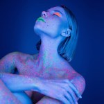 naked woman in bright makeup and neon body paint posing on dark blue background