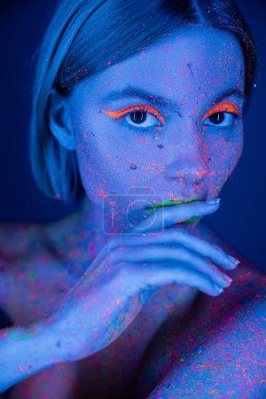 young woman with neon eye shadow touching lips and looking at camera isolated on dark blue