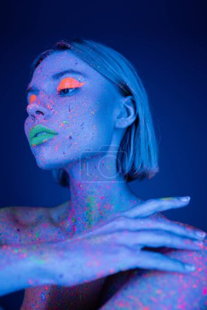 young woman with neon makeup touching body colored in fluorescent paint isolated on dark blue