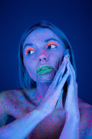 junge Frau mit leuchtendem Neon-Make-up und heller Farbe spritzt auf den Körper und schaut vereinzelt auf dunkelblauem Grund weg
