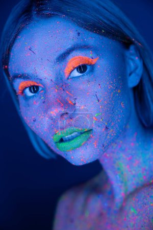 portrait of woman with glowing makeup and neon paint on face isolated on dark blue