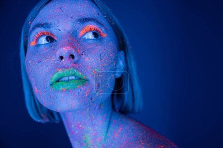 portrait of young woman with neon makeup and vibrant paint on face looking away isolated on dark blue
