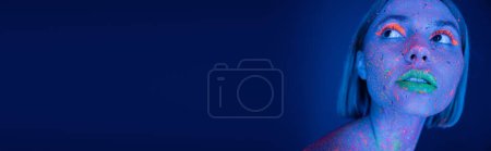 Téléchargez les photos : Portrait de jeune femme avec un maquillage néon vibrant regardant loin sur fond bleu foncé, bannière - en image libre de droit