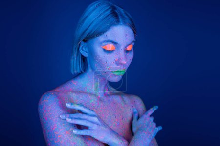 nackte Frau in hellem Neon-Make-up und glühender Körperfarbe, die Brust bedeckt mit Händen, die auf dunkelblau isoliert sind