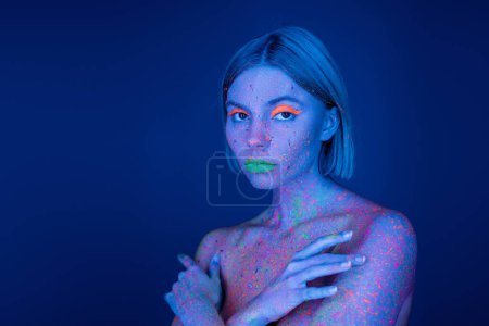 nackte Frau mit lebendigem Make-up und Körper in Neonfarbe, die isoliert auf dunkelblau in die Kamera schaut