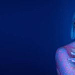 woman in glowing neon makeup and colorful paint splashes posing with crossed arms on dark blue background, banner