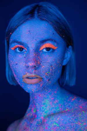 portrait of pretty woman with glowing makeup on face colored with neon paint looking at camera isolated on dark blue