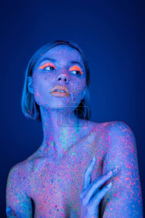 nackte Frau mit farbigem Körper und hellem Neon-Make-up, die isoliert auf dunkelblauem Grund wegschaut