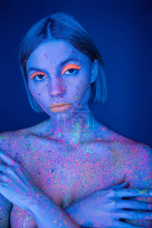 Foto de Mujer desnuda con maquillaje de neón y pintura brillante en el busto cubierta de cuerpo con las manos aisladas en azul oscuro - Imagen libre de derechos