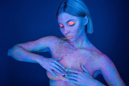 young woman in bright paint splashes and neon makeup covering breast with hands isolated on dark blue