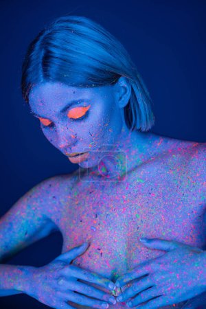 junge und nackte Frau mit leuchtendem Make-up und farbigem Körper, der Brust bedeckt, mit isolierten Händen auf dunkelblau