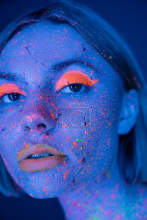 Téléchargez les photos : Portrait en gros plan de la femme en éclaboussures de peinture colorées et maquillage au néon isolé sur bleu foncé - en image libre de droit