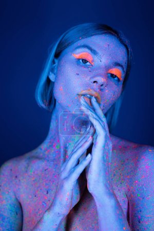 nackte Frau in leuchtendem Make-up und neonfarbener Körperfarbe, die Hände in Gesichtsnähe haltend, isoliert auf dunkelblau