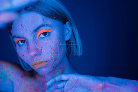 portrait of woman with fluorescent makeup and colorful neon body paint on blurred foreground isolated on dark blue