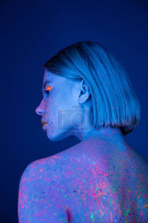 Rückansicht einer jungen Frau in leuchtendem Make-up und heller Neon-Körperfarbe isoliert auf dunkelblau