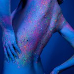 cropped view of naked woman in colorful neon paint posing isolated on dark blue