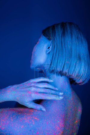 Rückansicht einer jungen Frau mit bunten Leuchtflecken auf dem Körper, die isoliert auf dunkelblauem Grund posiert