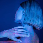 back view of young woman with colorful neon stains on body posing isolated on dark blue