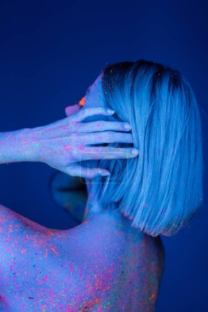 young woman colored with neon paint touching hair isolated on dark blue