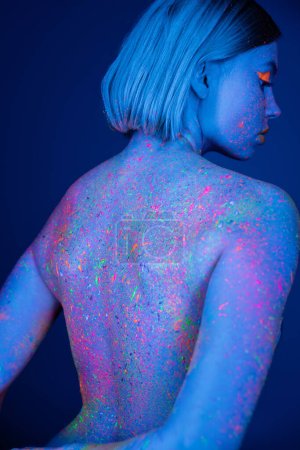 Téléchargez les photos : Vue arrière de la femme avec éclaboussures vibrantes de peinture au néon sur le corps isolé sur bleu foncé - en image libre de droit