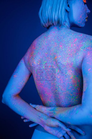 Frau mit Körper in hellen Neon-Spritzern, stehend mit den Händen auf dem Rücken isoliert auf dunkelblau