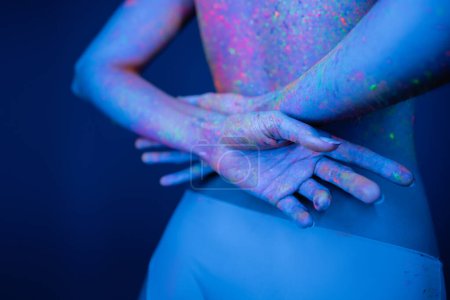 cropped view of young woman in bright neon paint holding hands behind back isolated on dark blue