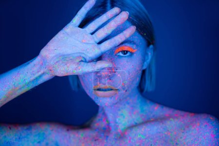 Photo for Woman in neon makeup and bright body paint obscuring face with hand while looking at camera isolated on dark blue - Royalty Free Image