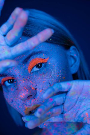 Téléchargez les photos : Femme au néon maquillage et éclaboussures de peinture brillantes posant avec les mains floues près du visage isolé sur bleu foncé - en image libre de droit